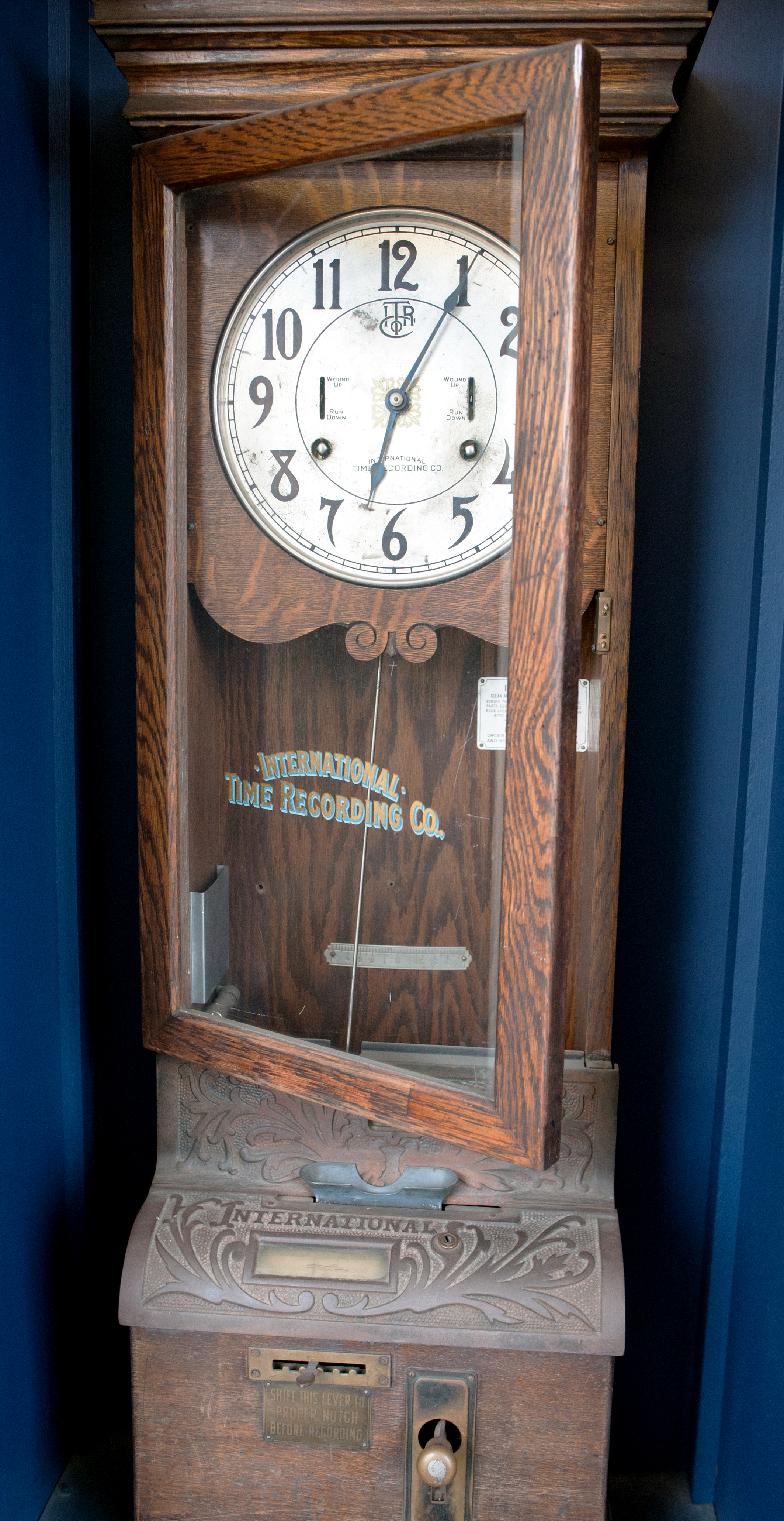 A wall mounted wooden cabinet clock that includes slots for tickets and a punch leaver. The lever must be shifted to record the time on the punch slips – the lever rests in the upright position.