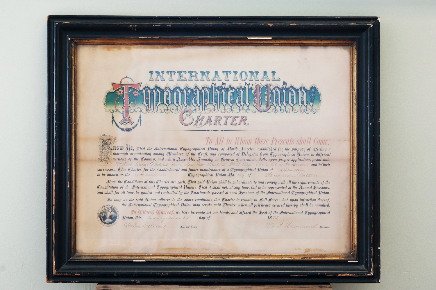 A photo of the International Typographical Union Charter from 1872. The signed paper charter uses historic calligraphy and has been preserved with glass and a wooden frame.