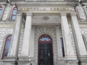 The entrance to a two-storey building. The door is framed by two pillars on each side and carved in the top of the portico entrance is the Latin 