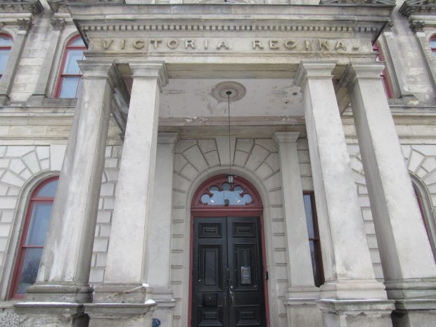 The entrance to a two-storey building. The door is framed by two pillars on each side and carved in the top of the portico entrance is the Latin Victoria Regina