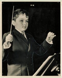 A young boy in a suit holds a conductor's baton in one hand and gestures with the other