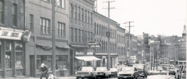 A streetscape of multi-storey brick buildings with stores on the bottom levels and cars from the late 1960s lining the street.
