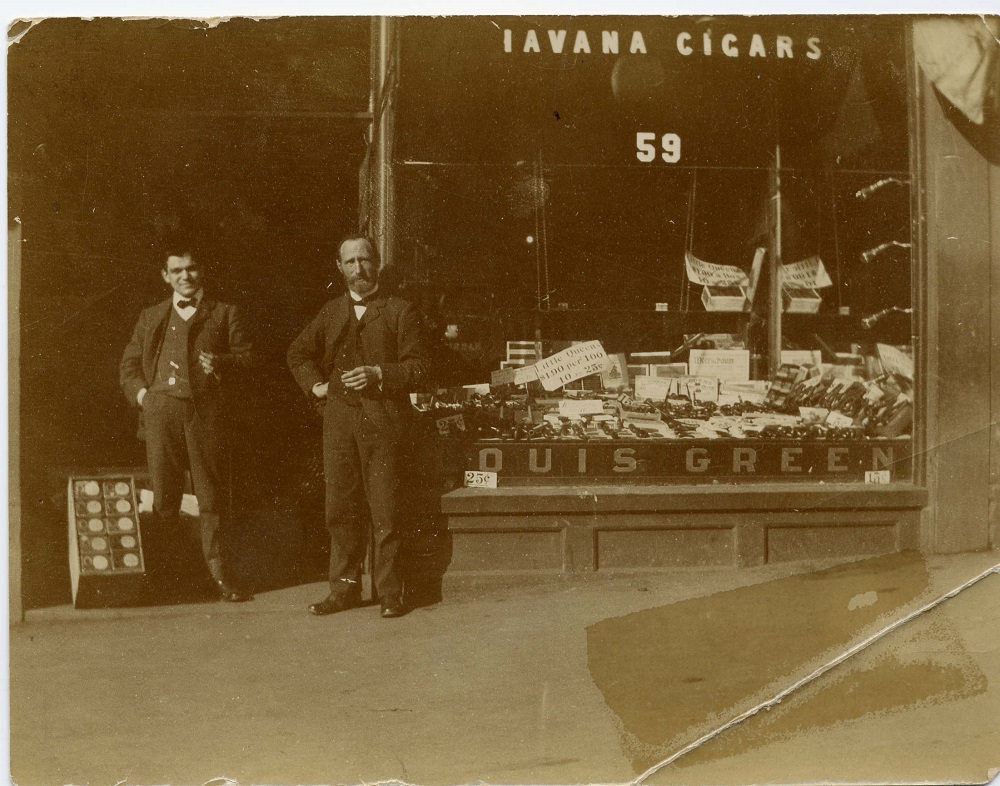 Storefront with large glass window with “Havana Cigars” written at the top of the window and “Louis Green” printed at the bottom – the window displays cigars and tobacco products – two men are standing in doorway.