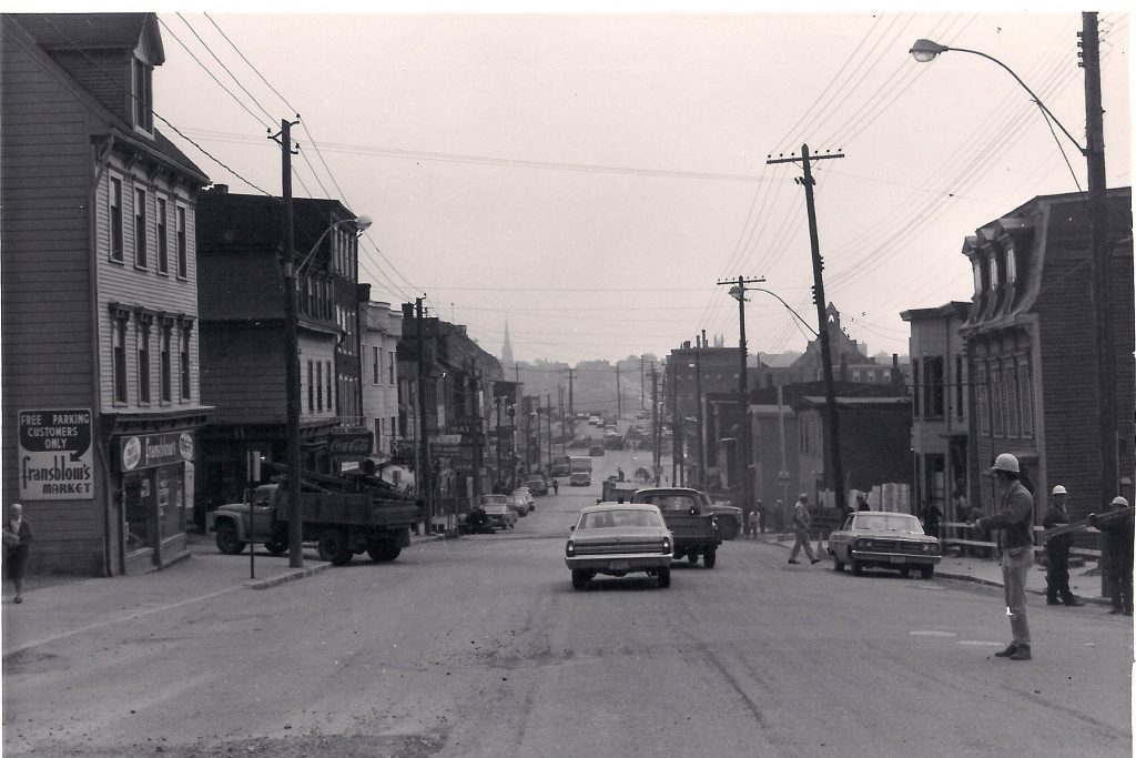 A view down the centre of Main Street with cars and trucks on the roadway and three-storey wooden buildings on either side, some with business signs including Fransblow’s Market.