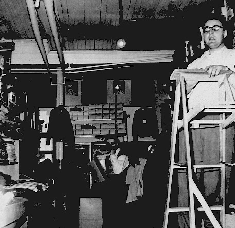 Interior of clothing store with shelving on the back wall for shoes and at left for folded shirts. One man is standing at the back of the store and another is on a ladder.