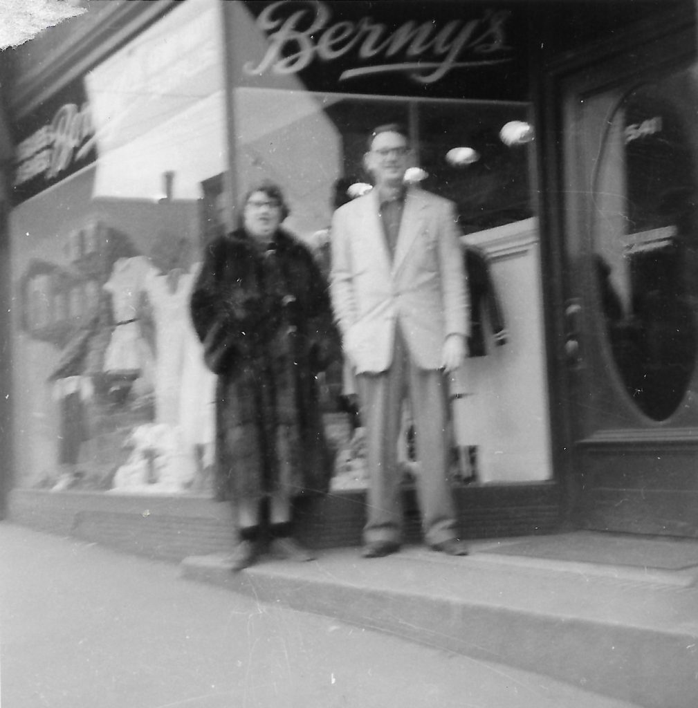 Un homme en costume et une femme en manteau de fourrure se tiennent à l’entrée d’un magasin où l’on peut lire « Berny’s » en lettres cursives sur la vitrine au-dessus de leurs têtes.