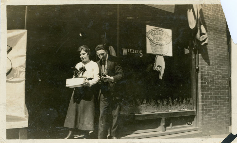 Store window with lettering for “Wiezel’s” – woman and man at left looking at a pair of shoes from a shoebox