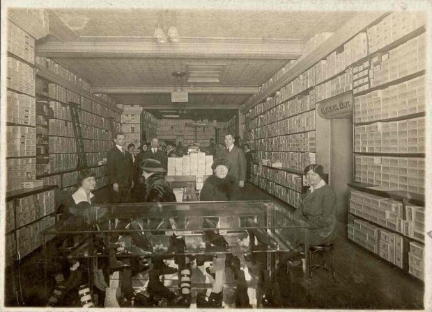 Interior of shoe store with boxes of shoes on shelves along walls on either side – glass table display at centre with female clerks and customers – men in background