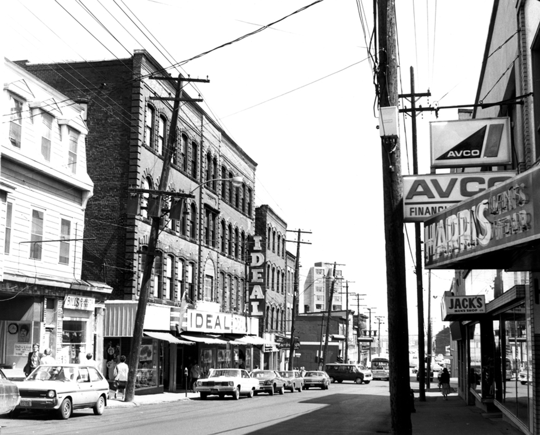 View of street lined with two and three storey buildings with stores at street level and signage including Bill’s Cigarette Centre, Ideal, Jack’s Men’s Shop and Harry’s Men’s Wear.