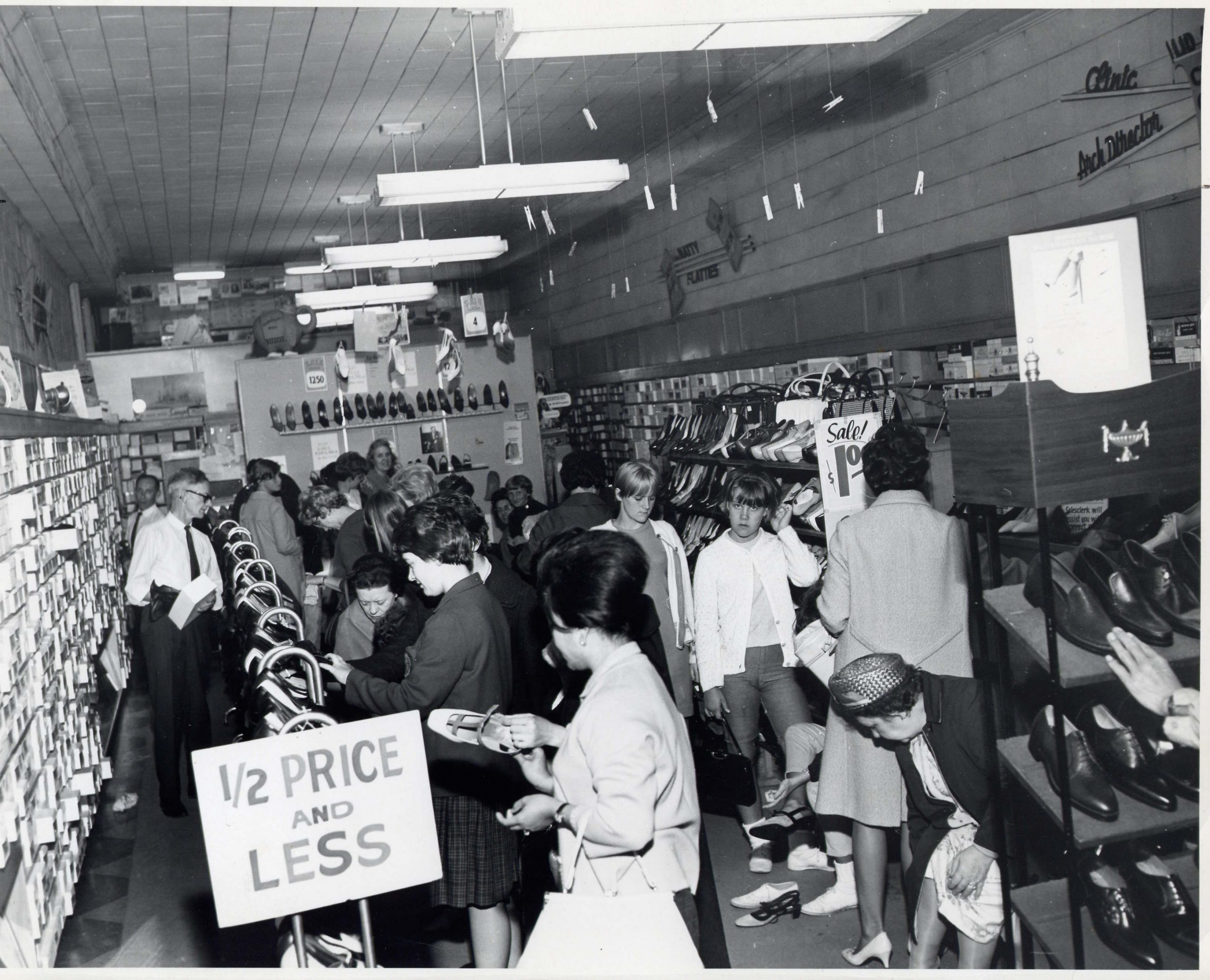 A narrow store crowded with women trying on shoes during a sale. Boxes and racks of shoes line the wall.