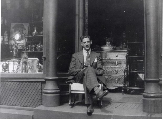 Man in suit seated in rocking chair in doorway to shop with wooden dresser and table in background. Shop window to left filled with objects.
