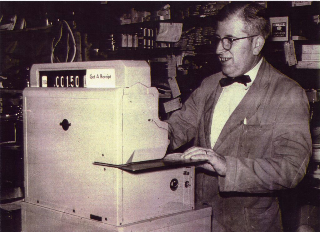 Man wearing round framed glasses and shop coat, standing in front of large cash register.