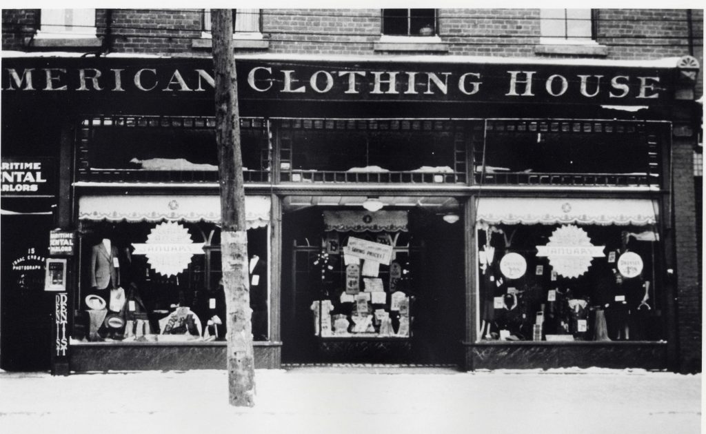 Magasin avec deux vitrines de chaque côté de l’entrée en retrait. Les vitrines présentent des costumes et des accessoires pour hommes. On peut lire American Clothing House au-dessus des fenêtres et de l’entrée.