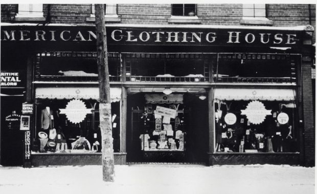 Store with two display windows on either side of recessed entrance – windows have a display of men’s suits and accessories. Lettering over windows and entrance reads American Clothing House.