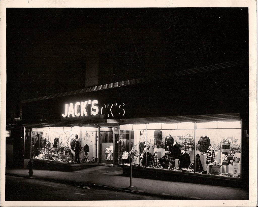 A night-time view of the store front of Jack’s Men’s Shop with large lighted glass display windows full of clothing displayed on mannequins or stands.