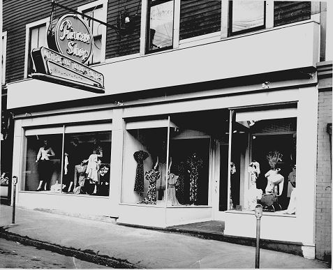 Wooden building with three large display windows with mannequins in dresses. Sign for Princess Shop hung on metal fitting over the door.