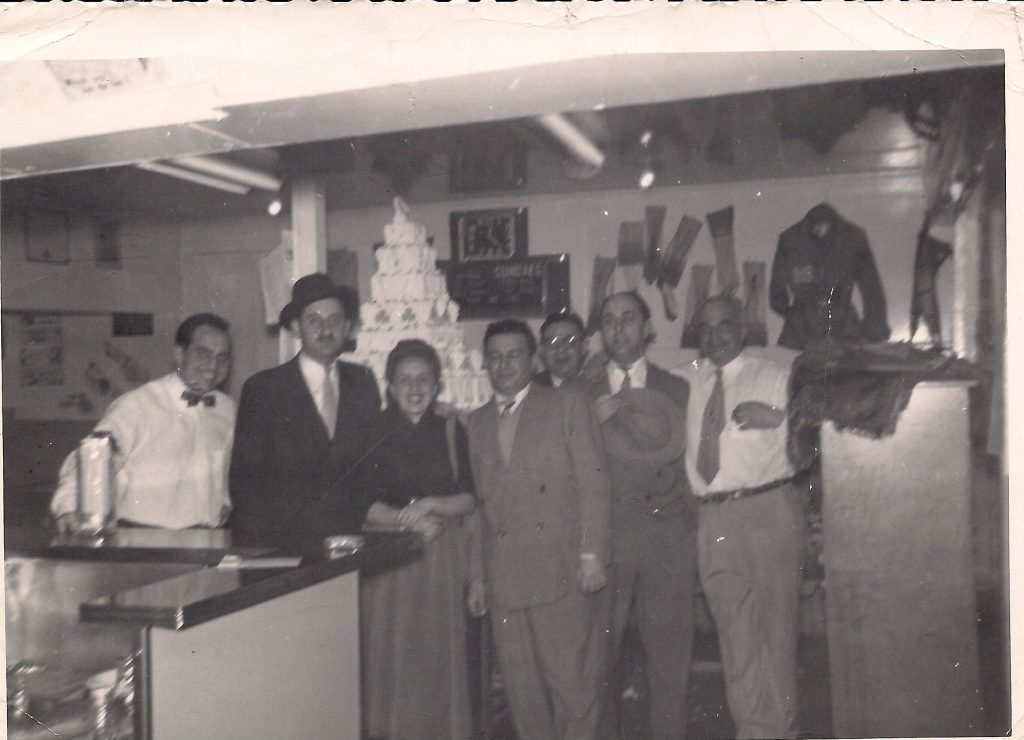 Interior of lunch counter and convenience store with group of six men and one woman standing at counter.