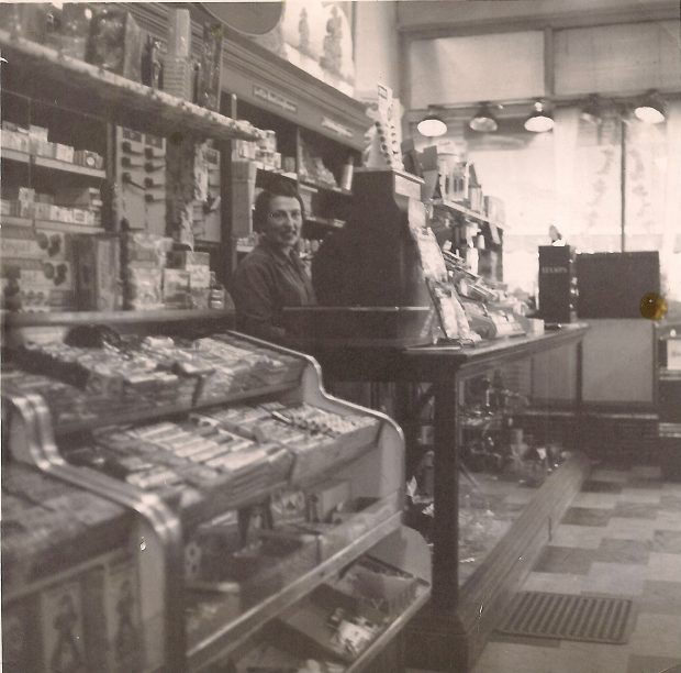 Interior of convenience store with counter filled with snack foods at left. Young woman standing behind large cash register.