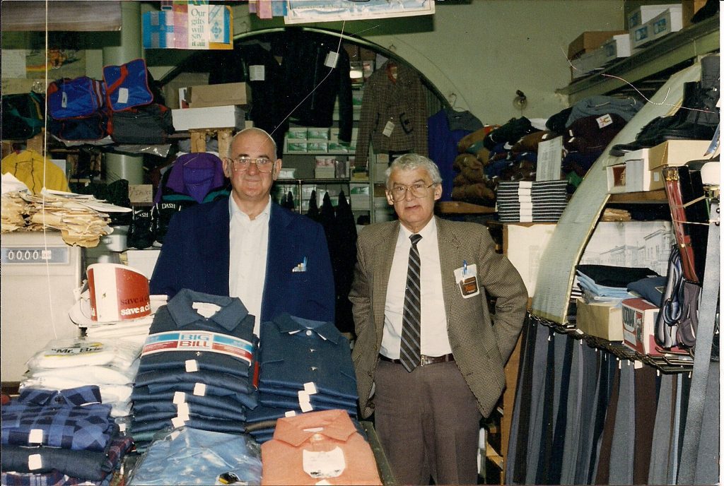 Two men inside cluttered clothing store and with piles of work shirts and flannel shirts in front of them.