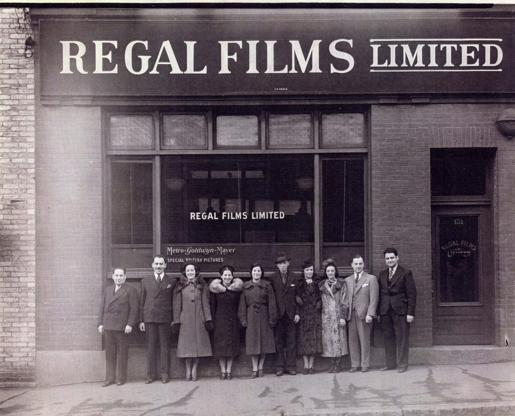 Groupe de cinq hommes et cinq femmes debout devant une fenêtre des bureaux de Regal Films Limited, dans un bâtiment en briques orné d’une grande fenêtre.
