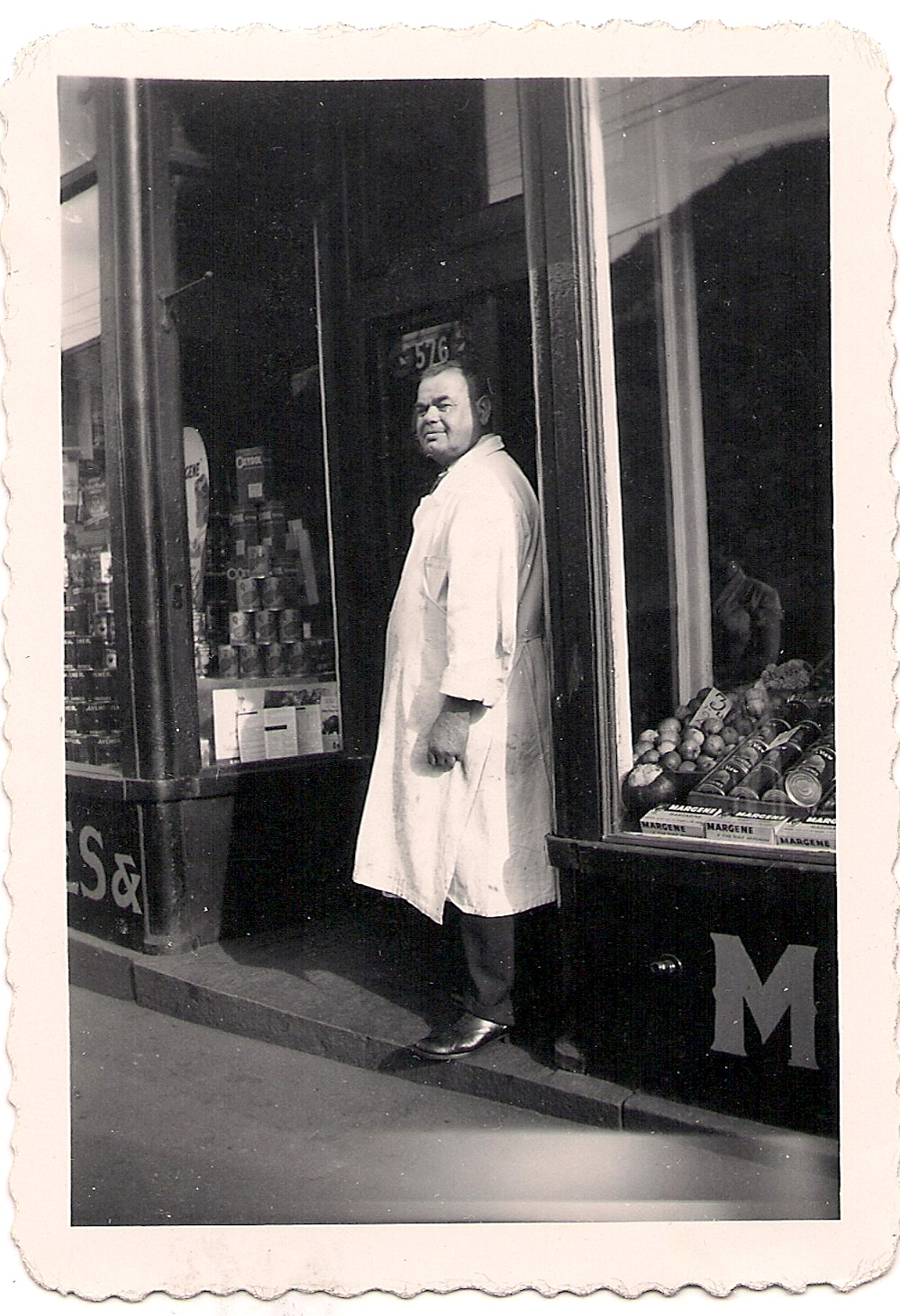 Man wearing long white coat standing at entrance to butcher shop - stack of canned goods in window.