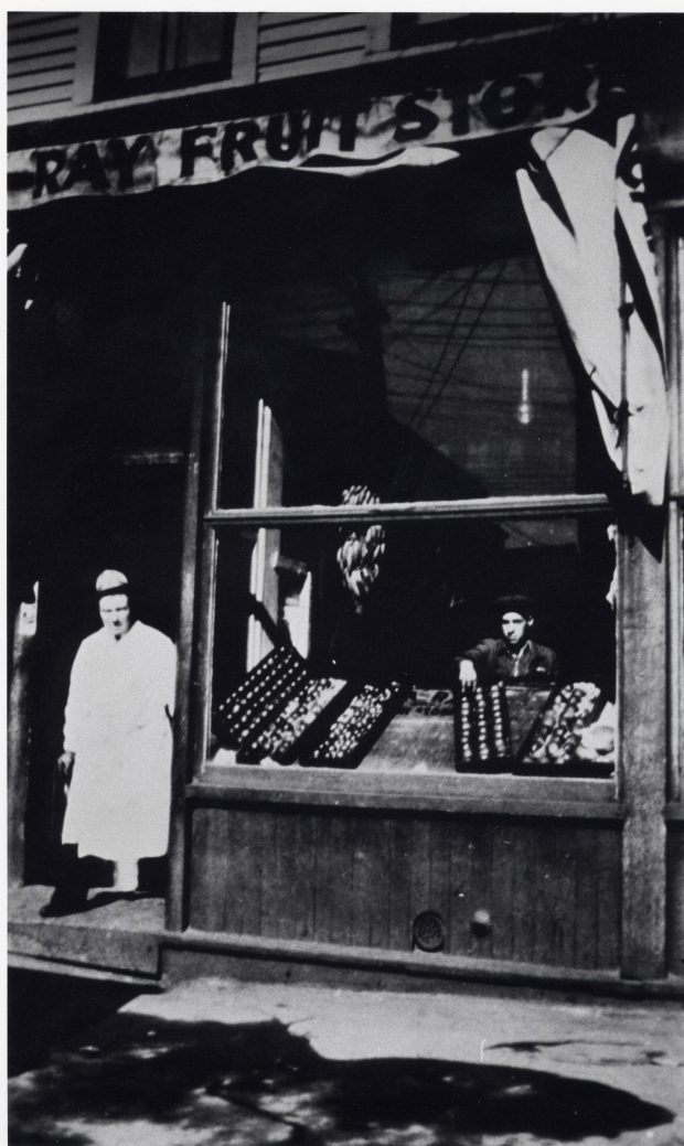 Man in a long work coat standing in the doorway next to a window displaying produce. Sun Ray Fruit Store written on a banner over the window.