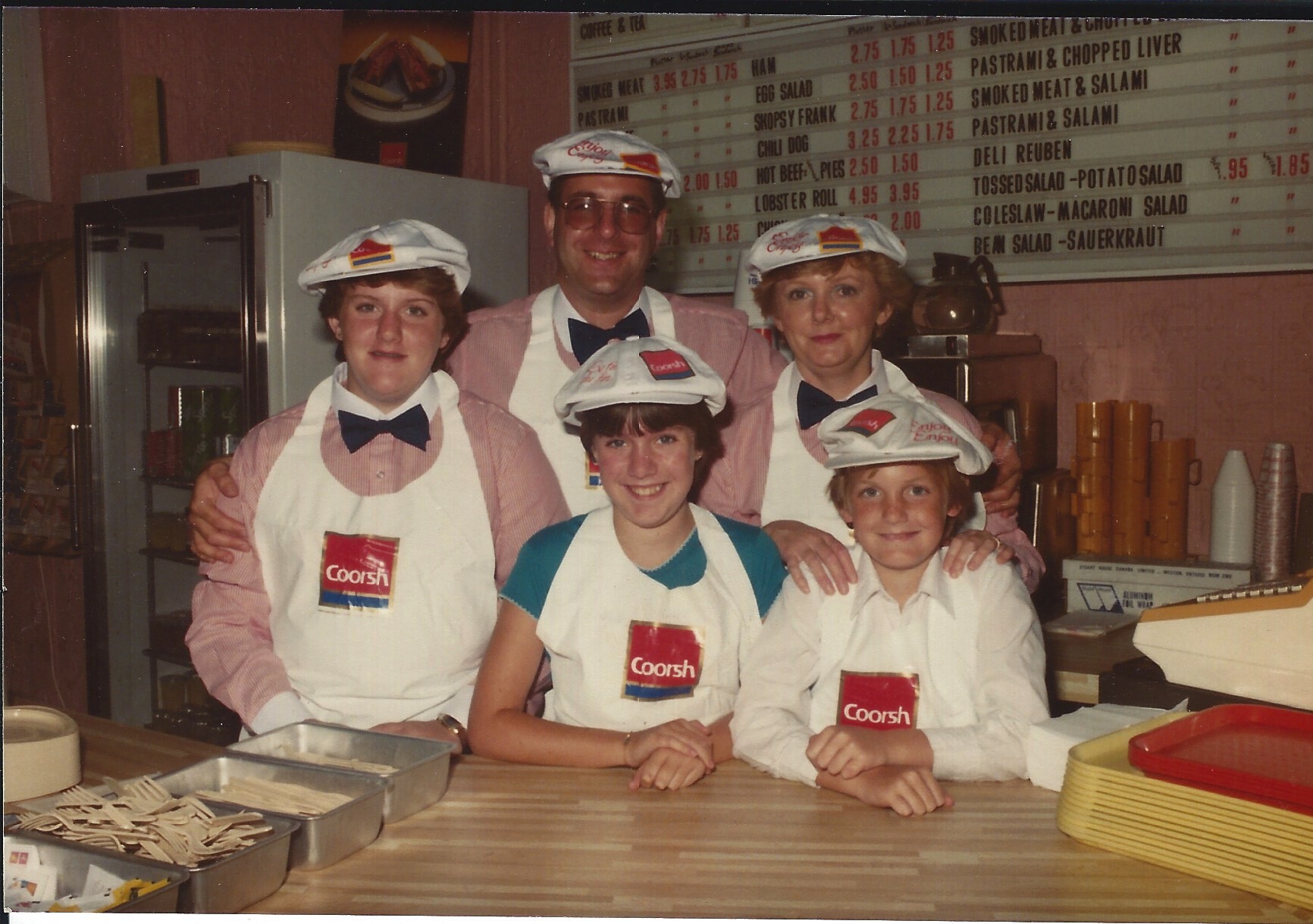 Man, woman and three teenagers standing behind lunch counter. Menu on the wall behind them.