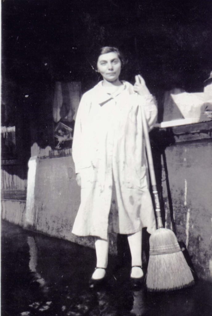 Une femme aux cheveux courts, portant un manteau long, tient un balai de paille dans la main gauche.