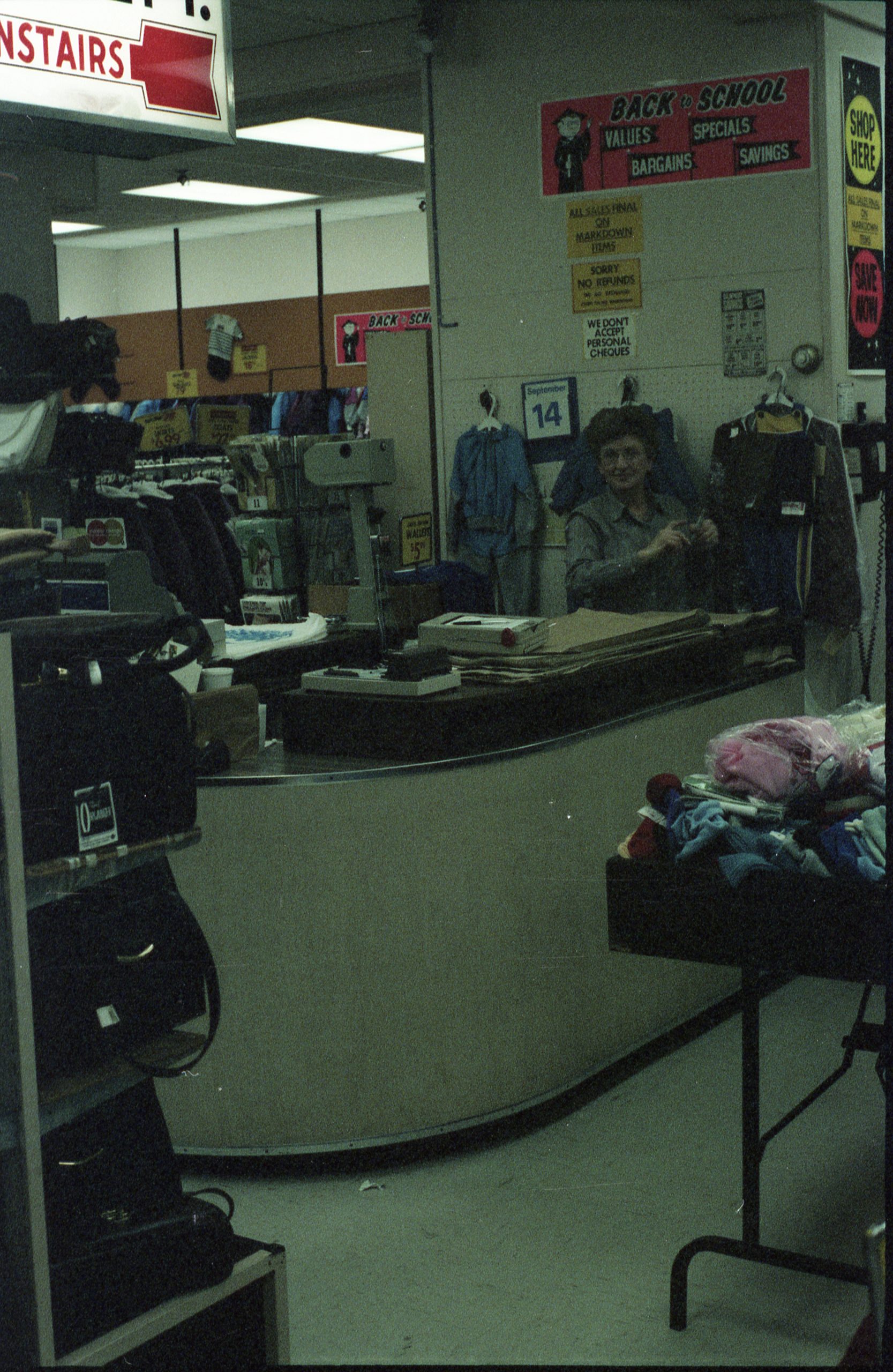 Woman standing behind a curved counter – stands with handbags and clothing in foreground.