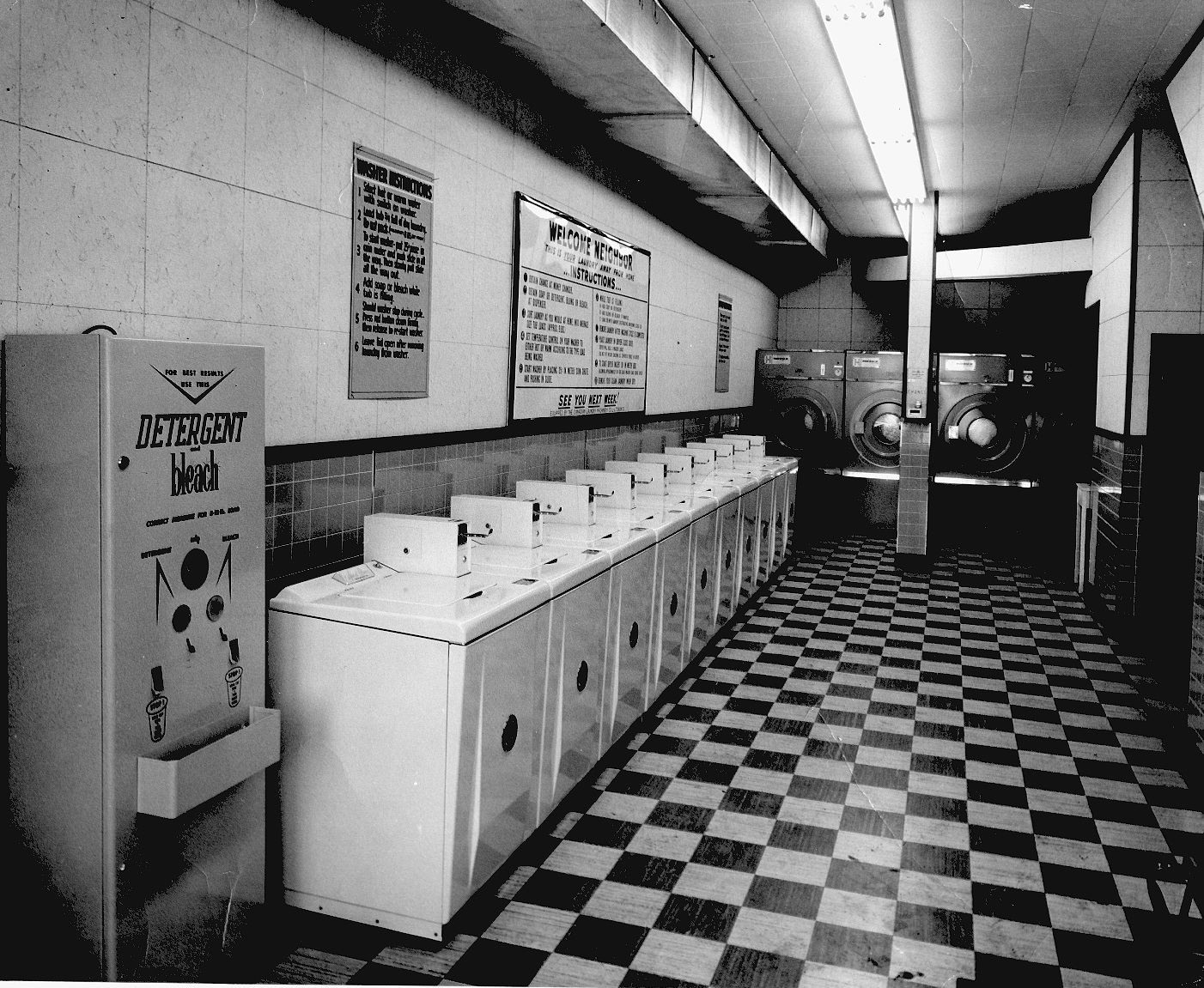 A row of ten washing machines and a laundry soap dispenser. Large clothes dryers on back wall.