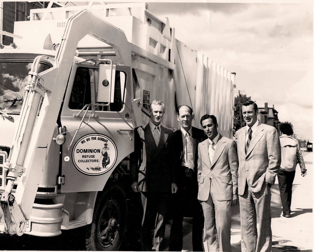 Four men standing alongside a large truck with front arm for emptying dumpsters. The sign on the driver’s door reads “We Go For Garbage – Dominion refuse Collectors” with a design of a cat pushing a broom.