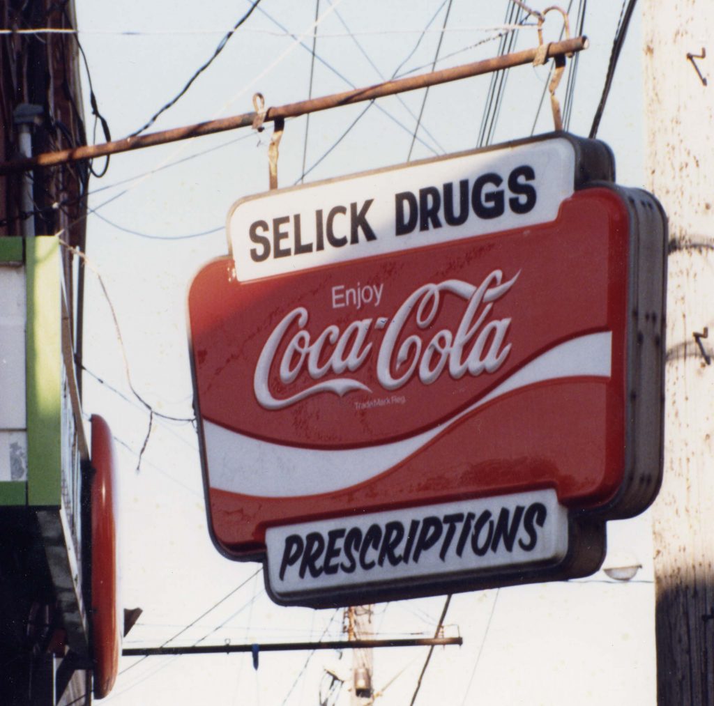 Enseigne accrochée à un support métallique sur le côté d’un bâtiment, où on lit « Selick’s Drugs » et « Prescriptions » avec un grand logo Coca-Cola entre les deux.