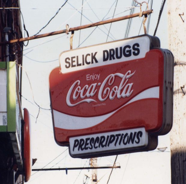 Sign hanging on metal bracket on side of building – Selick’s Drugs” and “Prescriptions” with a large Coca-Cola logo in between.