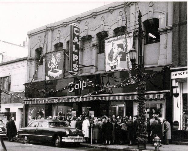 Exterior view of Calp’s Store with large signs over the entrance, Christmas decorations and a large line of customers outside waiting to enter.