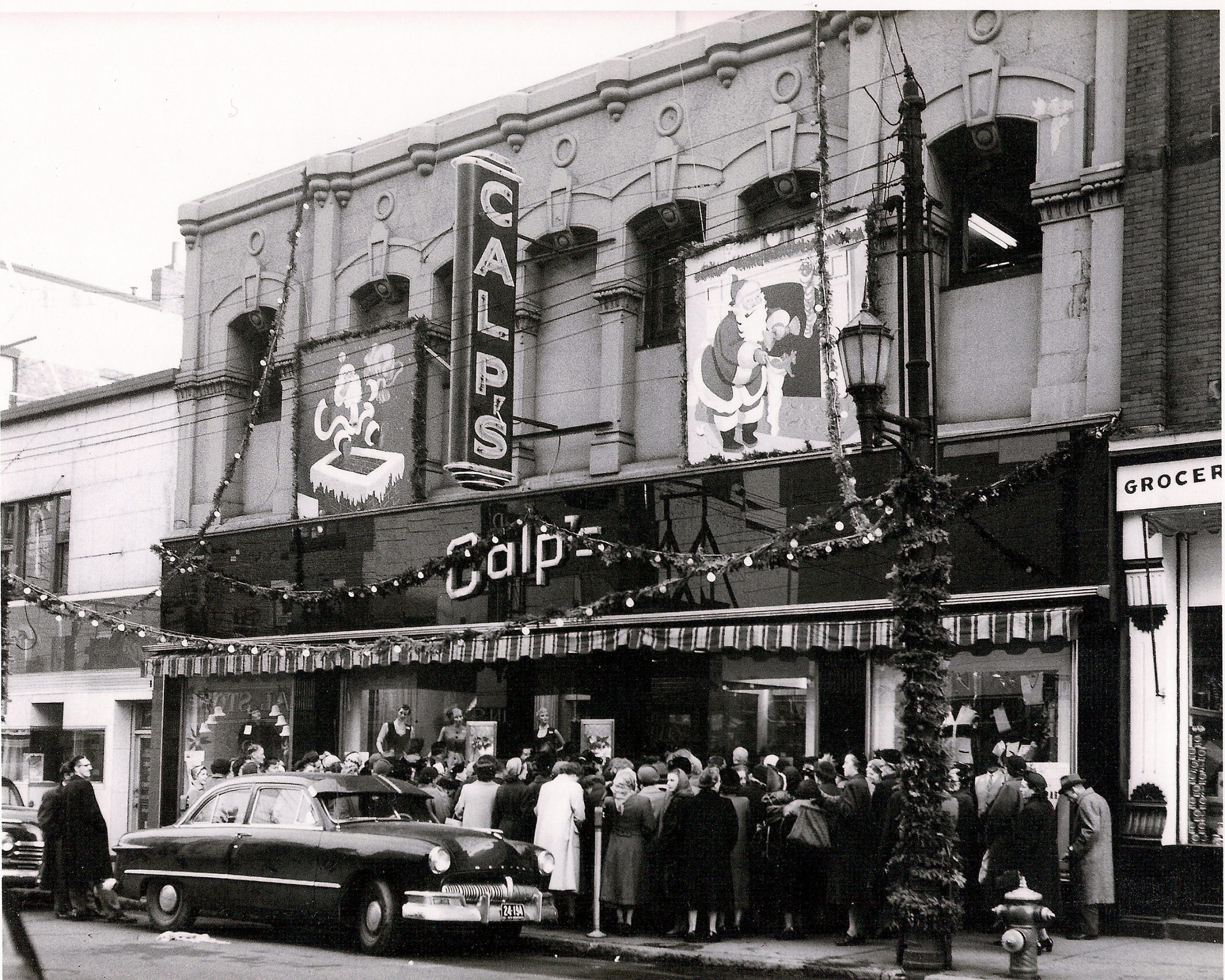 Exterior view of Calp’s Store with large signs over the entrance, Christmas decorations and a large line of customers outside waiting to enter.