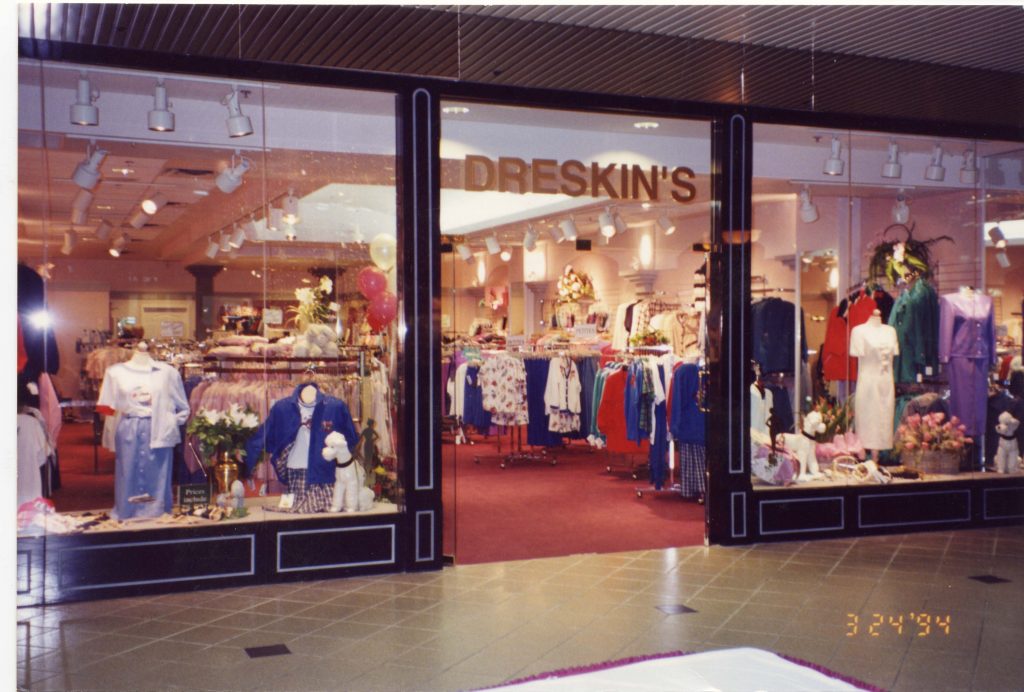 Entrée d’un magasin de vêtements pour dames avec des fenêtres entièrement vitrées et une large entrée. Des étagères de blouses et de robes pour femmes sont visibles à l’intérieur du magasin. Le nom « Dreskin’s » est imprimé en lettres dorées au-dessus de l’entrée.