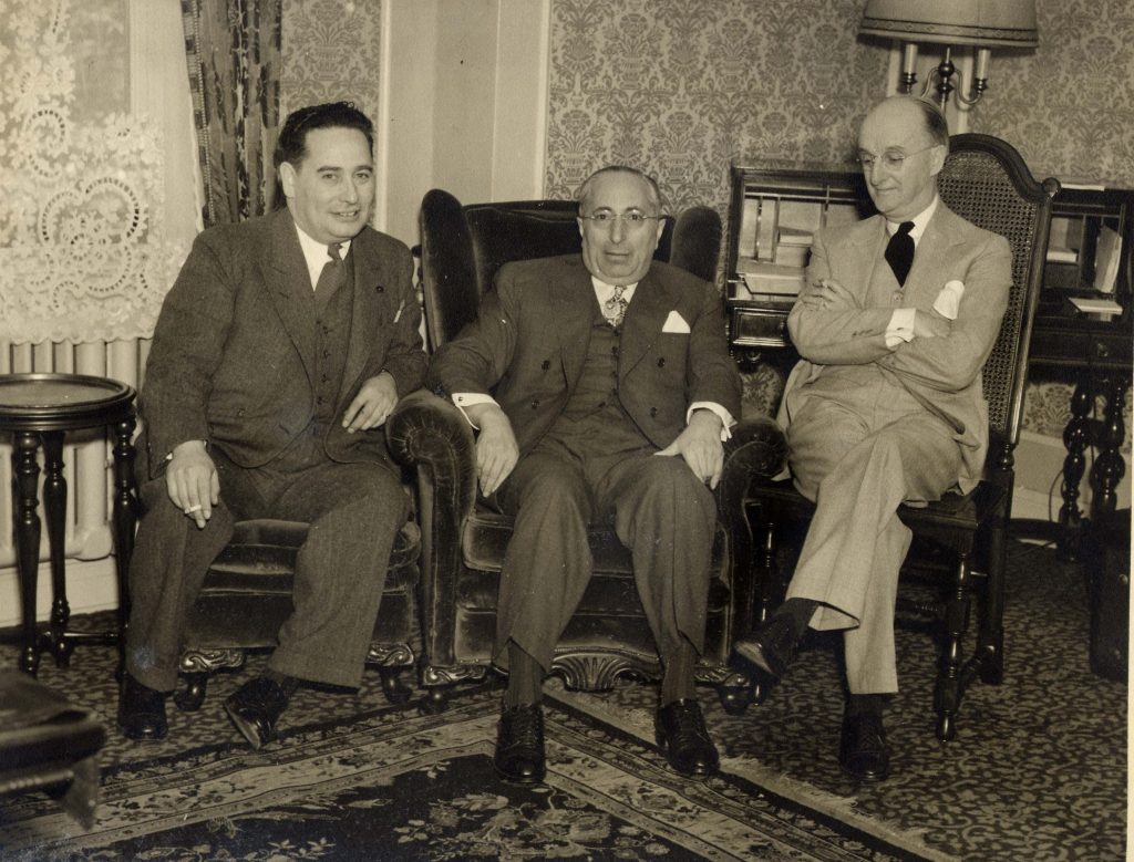 Three men dressed in suits are seated in arm chairs.