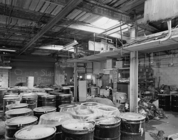 Black and white photograph depicting an industrial storage room. It is a large, disordered room clearly left abandoned. It is filled with metal barrels of unknown content.