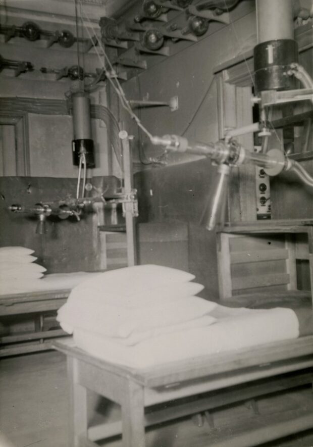 Black and white photograph depicting the corner of a room filled with machines and wiring. Two devices composed of several interconnected tubes suspended from the ceiling and attached to the walls are installed above two tables equipped with mattresses and pillows.