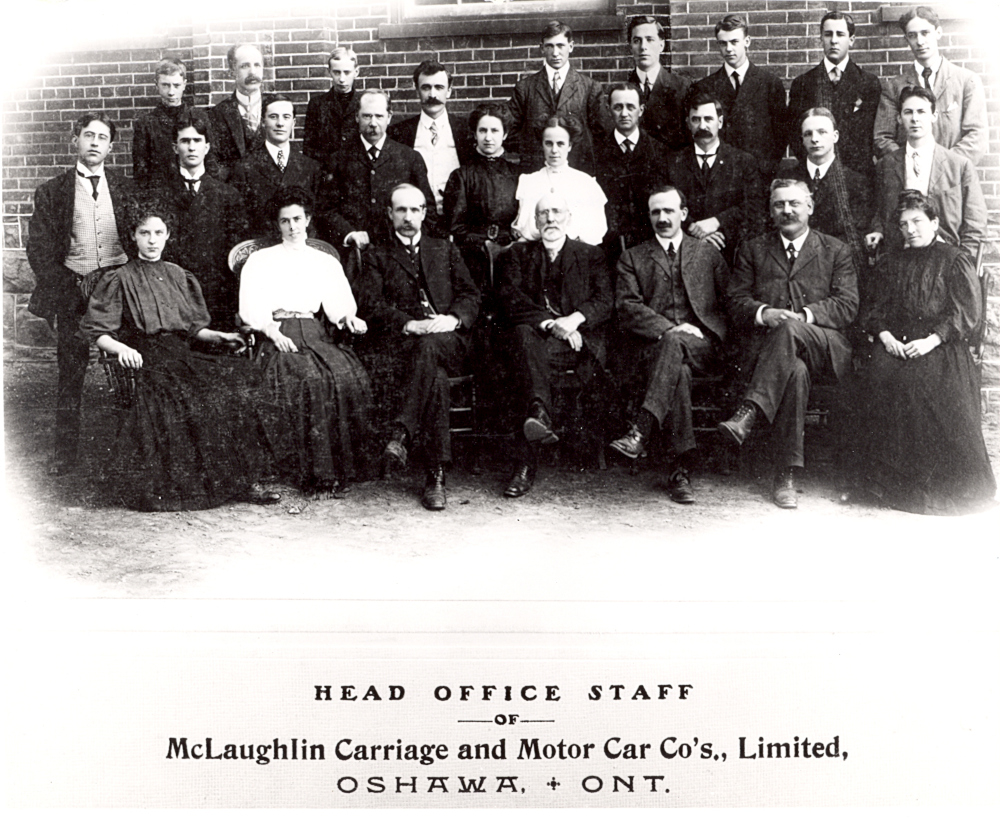 Black and white image of several dozen male and female office workers in formal 1900s clothing posed in front of a brick wall. Caption reads "HEAD OFFICE STAFF OF McLaughlin Carriage and Motor Car Co's., Limited, OSHAWA, + ONT"