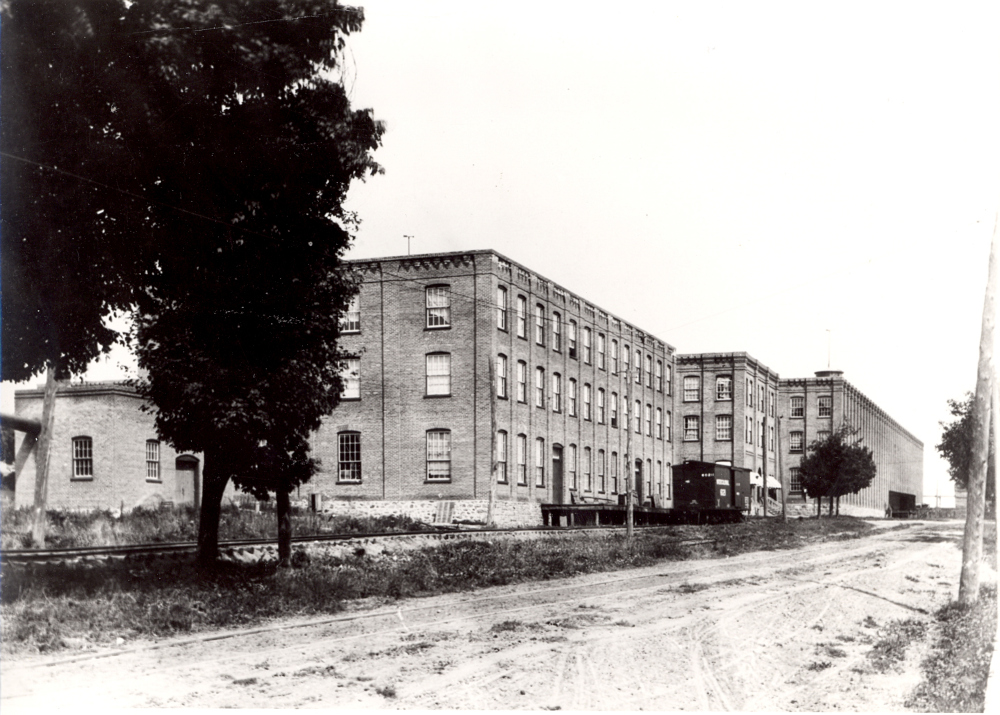 An exterior view of a set of large factory buildings. A railway line with a box car parked on it runs in front of the buildings.