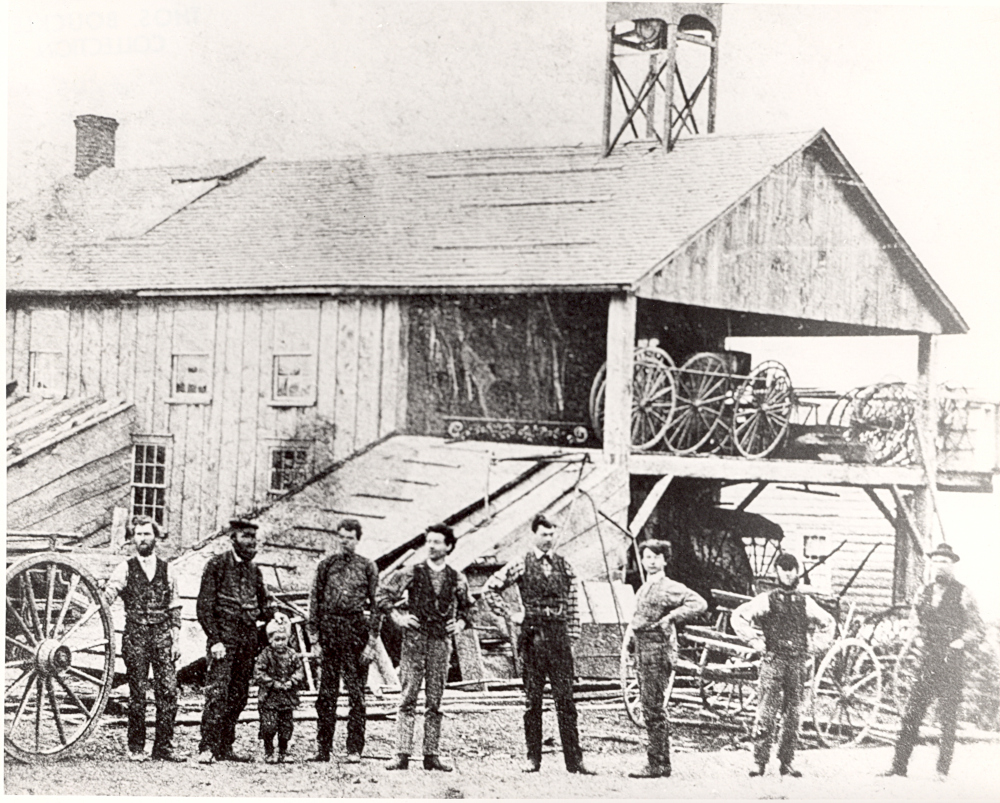 Black and white image of eight men and a young boy posed in a line outside a wooden building, surrounded by carriages and carriage parts.
