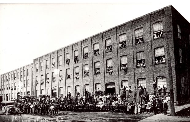 Black and white image of several hundred factory workers posing with four automobiles in front of a large factory building.