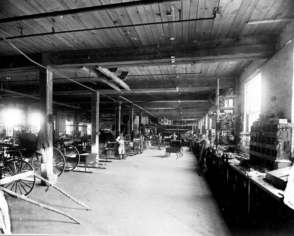 A factory room. Carriages and sleds are spread through the space. Several workers are standing in front of a tool bench.