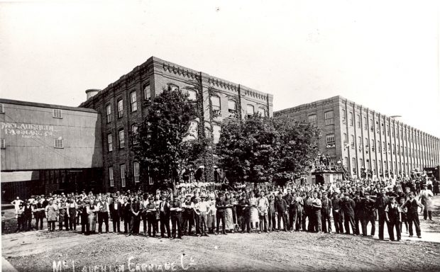 A crowd of several hundred employees pose in front of a series of large factory buildings.