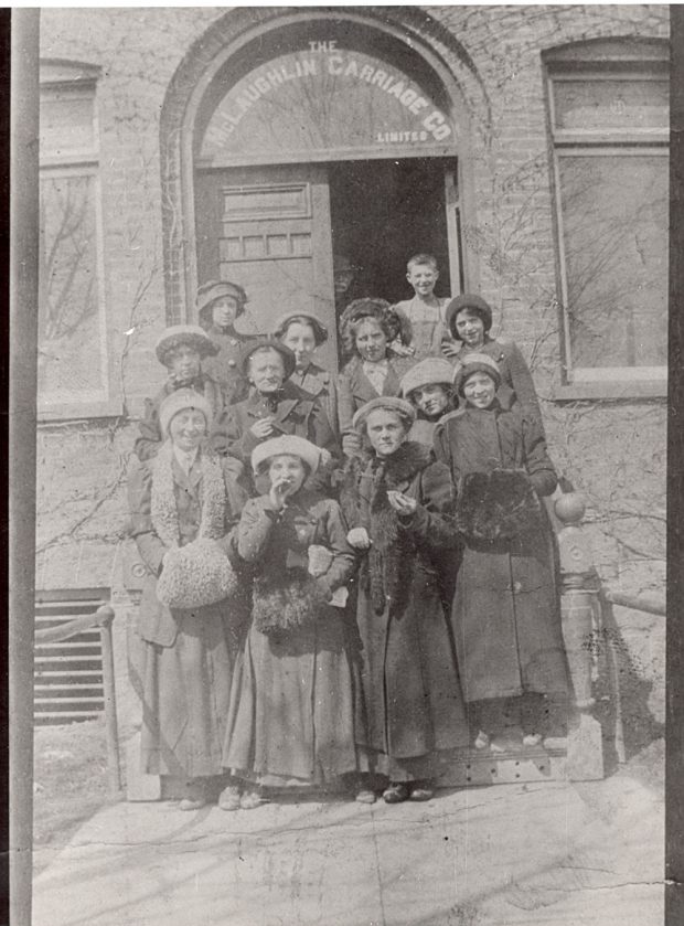 Female office workers in 1900s clothing stand on the steps of a brick building with The McLaughlin Carriage Co Limited printed above the doorway.