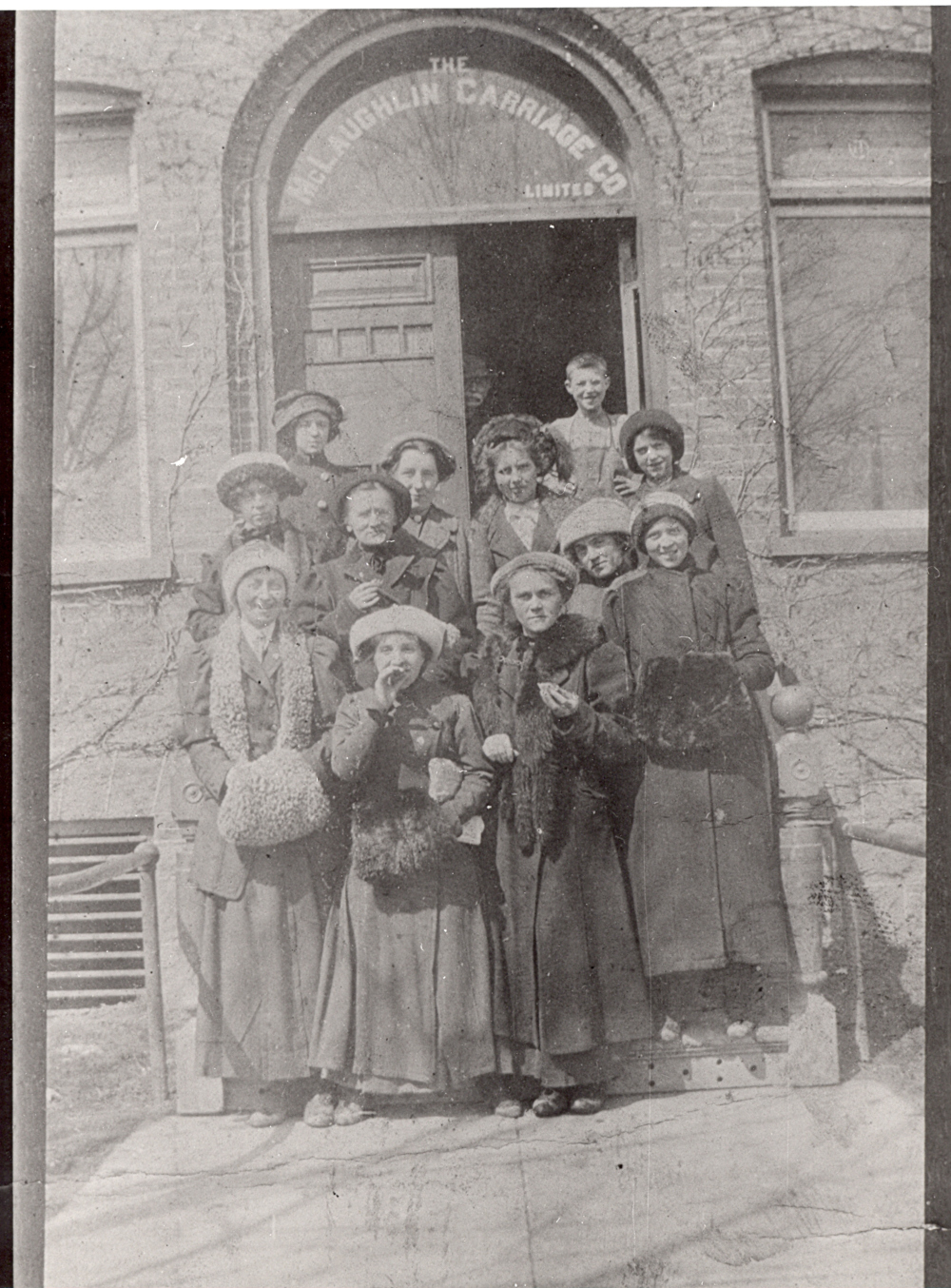 Female office workers in 1900s clothing stand on the steps of a brick building with "The McLaughlin Carriage Co Limited" printed above the doorway.