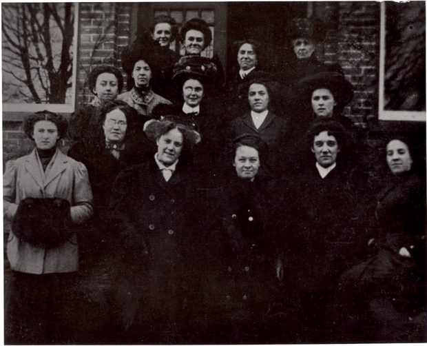 Fifteen female office workers in formal 1900s clothing stand on the steps of a brick building.