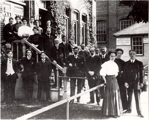A group of several dozen male and female office workers in 1900s formal wear stands on the steps of a building overgrown with ivy.