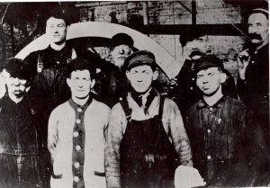 A group of eight factory workers stand in front of the wheel of a large piece of machinery.